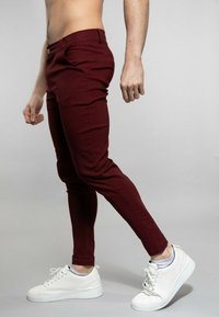 Pantalons skinny bordeaux avec poches avant et fermeture à bouton. Portés avec des baskets blanches, mettant en avant un design ajusté et une texture lisse.
