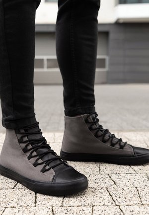 Scarpe da ginnastica alte con tomaia in tela grigia, punta in gomma nera e sistema di allacciatura. Superficie texturizzata e costruzione robusta.
