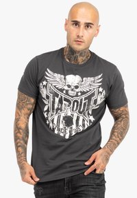 Graues Kurzarm-T-Shirt mit einem weißen Totenkopf-Design und Flügeln sowie dem Text "TAPOUT" in fetter Schrift, vor einem strukturierten Hintergrund.