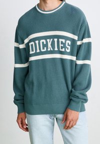 Zelenomodrý pletený svetr s bílými pruhy a nápisem "DICKIES" přes hruď. Žebrované manžety a lem dodávají texturu a zajišťují pohodlné nošení.