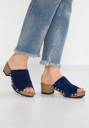 Fødder iført blå ruskind åbne træsko med træklodshæle og sølvnitter, kombineret med lyseblå denimjeans med flossede kanter.