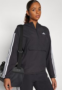 Svart lett pullover-jakke med hette, med hvite vertikale striper på ermene, en frontlomme og glidelås. Svart treningsbag inkludert.