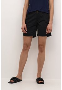 Pantalones cortos de algodón negros con dobladillos enrollados, diseño de cinco bolsillos y hardware de latón. Llevados con sandalias negras tipo slide.
