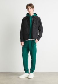 Svart lett jakke med to frontlommer over en grønn hettegenser, kombinert med grønne joggebukser og hvite sneakers på en nøytral bakgrunn.