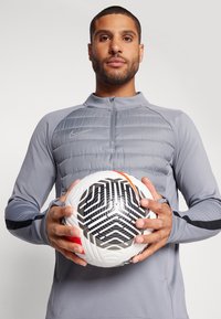 Mann in grauem Nike-Sportanzug, der einen weißen Fußball mit schwarz-rot gemustertem Design hält, frontal vor einem einfarbigen Hintergrund.