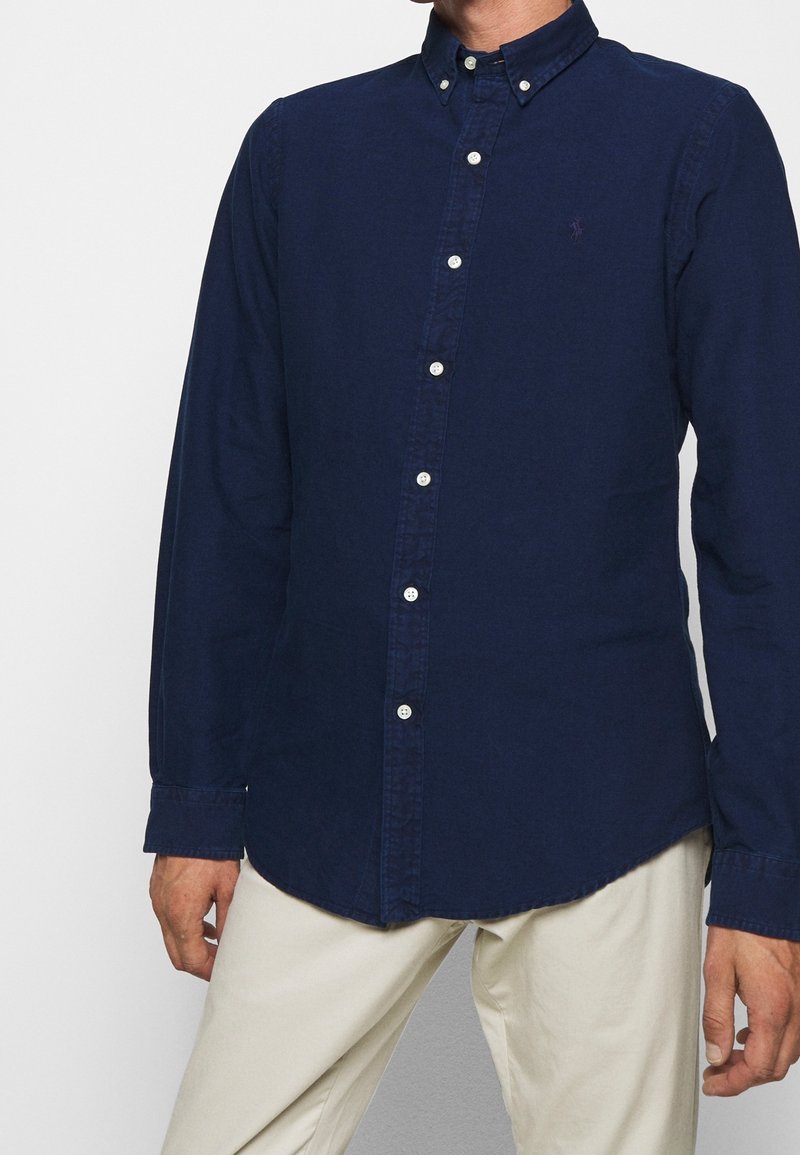 Marinblå knapp-up skjorta med spetsig krage, långa ärmar och vita knappar. Har varumärkeslogotyp på bröstet. Slät bomullstyg.