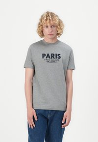 Pilkas medvilninis marškinėlis su ryškiu tamsiai mėlynu "PARIS" spaudiniu priekyje. Apvalus iškirpimas ir trumpomis rankovėmis. Dėvimi su mėlynomis džinsinėmis kelnėmis.
