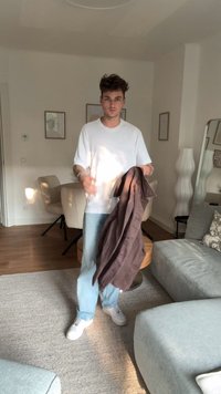 eller klær med en hvit t-skjorte og vide blå jeans. Holder en brun, strukturert jakke. Bakgrunnen inkluderer en lysfarget lenestol og dekor.
