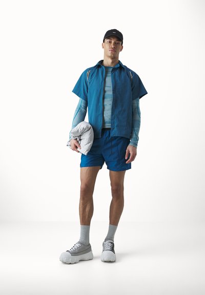 Športový outfit pozostávajúci z modrej tričkaj s krátkym rukávom, vzorovanej bundy s dlhým rukávom, modrých šortkách, šedých teniskách a čiernej šiltovke. Drží šedú bundu.