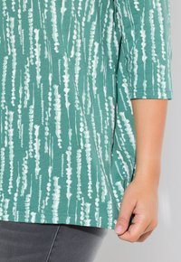 T-Shirt in Türkis mit weißen abstrakten vertikalen Streifen, drei Viertel Ärmeln und lockerer Passform. Glatte Textur mit abgerundetem Saum.