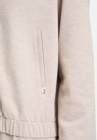 Jasno beżowa kurtka o teksturowanej tkaninie, z boczną kieszenią i złotym zatrzaskiem z przodu. Posiada elastyczny pas.