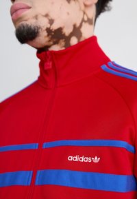 Červená sportovní bunda na zip s modrými horizontálními pruhy, logem adidas na levé straně hrudníku, hladkým materiálem a vysokým límcem.