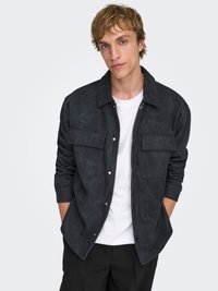 Veste en daim gris foncé avec deux poches poitrine, fermeture par boutons-pression, manches longues portée sur un t-shirt blanc, associée à un pantalon noir.