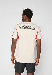 Šviesiai pilkas sportinis džersis su raudonomis detalėmis, trumpomis rankovėmis, turi juodą "SAGRES" logotipą nugaros pusėje, lygus tekstūra ir sportiškas pjūvis.