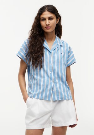Frau mit langen lockigen Haaren, die ein blau-weiß vertikal gestreiftes Kurzarm-Hemd mit Knöpfen und weiße Shorts trägt, Hände in den Taschen.