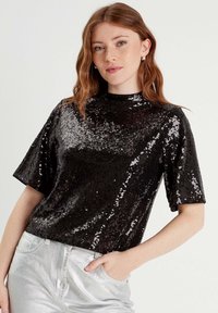 Top corto de lentejuelas negras con diseño de cuello alto, que presenta una textura brillante, combinado con pantalones metálicos plateados.