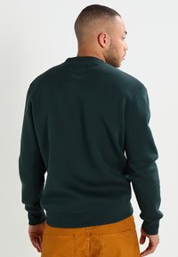 Sötétzöld pulóver, kerek nyakkal, bordázott mandzsettákkal és derékpánttal. Simas texture, kis hátsó címke részlettel.