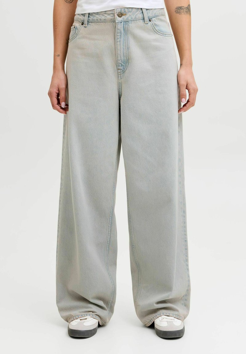 Jean large coupe délavé clair à taille haute avec fermeture à boutons, porté avec des baskets blanches et un haut blanc à manches courtes.