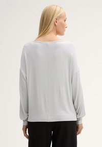 Hellgraues, lockeres Top mit weitem Ausschnitt, langen puffigen Ärmeln und glatter Textur. Eine Naht verläuft senkrecht über den Rücken.