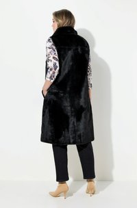 Gilet in pelliccia sintetica nera con colletto alto, design alla lunghezza del ginocchio e tasche laterali, indossato sopra una camicia a fantasia e abbinato a jeans scuri e tacchi nudi.