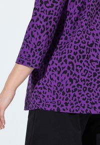Lila Oberteil mit einem schwarzen Leopardenmuster. Verfügt über Dreiviertelärmel und eine lockere Passform, kombiniert mit schwarzen Hosen.