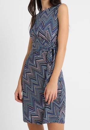 Mouwloze jurk tot op de knie met een zigzagpatroon in blauw, zwart en wit, voorzien van een strik aan de zijkant bij de taille.