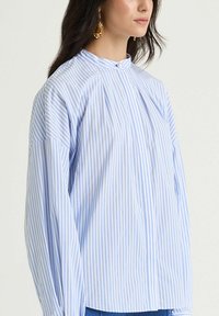 Camicia a maniche lunghe con righe azzurro chiaro e bianche, collo alto, con pieghe e bottoni centrali, realizzata in tessuto di cotone.