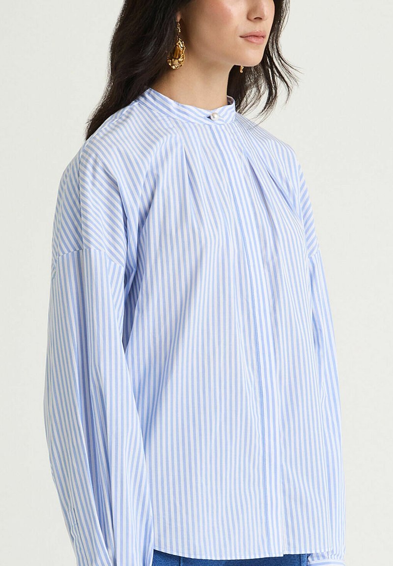 Camicia a maniche lunghe con righe azzurro chiaro e bianche, collo alto, con pieghe e bottoni centrali, realizzata in tessuto di cotone.