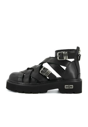 Zwarte chunky platform sandaal met gekruiste bandjes, twee zilveren gespen en een metalen merkplaatje aan de buitenzijde van de zool.