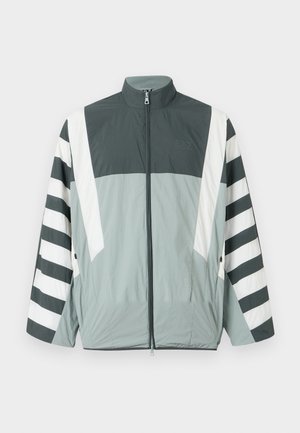 Lett zip-up jakke har et grått og hvitt fargeblokkdesign med markerte striper på ermene. Laget av glatt, slitesterkt stoff.