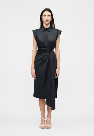 Kvinde stående iført en ærmeløs sort midi-kjole med skulderpuder, bælte i taljen med draperet stof og hvide stropper på hælene.