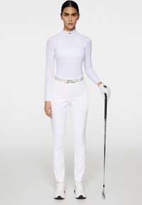 Biała koszula z długim rękawem o wysokim kołnierzu, połączona z białymi spodniami i beżowym paskiem. Modelka ma na sobie szary rękawiczki i stoi z kijem golfowym.