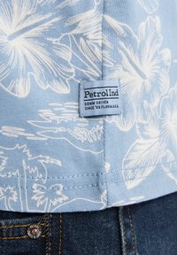 Svetle modrá košile s bílými květinovými vzory, s cedulkou označenou "PetrolInd" na spodním lemu, nošená přes tmavé džíny.