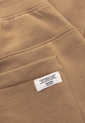 Gros plan sur une poche en tissu marron avec une étiquette blanche portant l'inscription « YOUR ONLY LIMIT IS YOUR MIND » et « OMBRE since 1985 » cousue sur la poche.