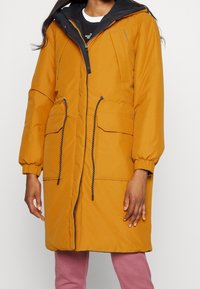 Parka aislante de color mostaza con cierre frontal de cremallera, puños elásticos, dos bolsillos frontales y capucha forrada en negro. Cuenta con cordones ajustables en la cintura.