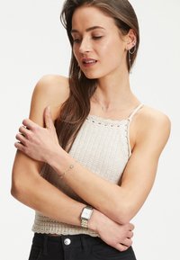 Silberne Uhr mit rechteckigem Zifferblatt und farblichen Akzenten, kombiniert mit einem filigranen Armband, das runde und ovale Charms enthält, getragen über einem gestrickten beigen Oberteil.