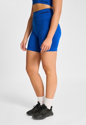 Kobieta w niebieskich bezszwowych szortach marki hummel, dopasowanym sportowym biustonoszu, białych skarpetkach i czarnych butach sportowych na jednolitym tle.