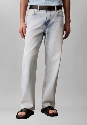 Mand iført lysvaskede straight-leg jeans, sort bælte, hvid skjorte og sorte slidesandaler, stående mod en ensfarvet grå baggrund.