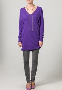 Fialový oversized svetr s výstřihem do V, v kombinaci se šedými úzkými kalhotami a fialovými lodičkami na podpatku. Doplněno stříbrným náhrdelníkem.