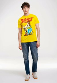 Yellow cotton T-shirt featuring a graphic print with characters from Asterix. Model pairs it with blue jeans and beige sneakers.