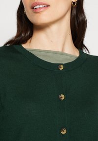 Ciemnozielony kardigan o prążkowanej fakturze, z okrągłym dekoltem i dwoma brązowymi guzikami. Pod spodem widoczna oliwkowozielona bluzka.