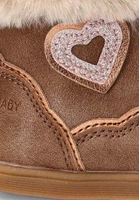 Zapato marrón con textura suave, que presenta un ribete de piel y un acento en forma de corazón rosa brillante. Los detalles cosidos realzan el diseño general.