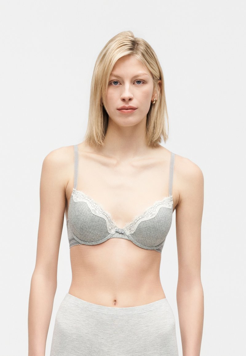 Ung kvinde med glat blondt hår iført en grå bh med blonder og matchende lysegrå underdele, stående mod en simpel hvid baggrund.