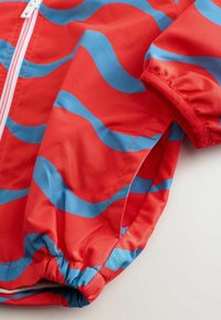 Tissu rouge avec des rayures bleues irrégulières, poignet élastique et couture de fermeture éclair blanche sur un vêtement.