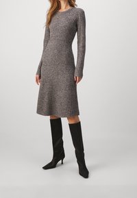 Robe tricotée côtelée grise avec des manches longues, ourlet jusqu'au genou, encolure ronde, portée avec des bottes noires à talons hauts jusqu'aux genoux. Texture lisse, silhouette cintrée.