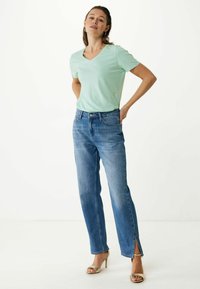 Světle zelené tričko s výstřihem do V, spárované s modrými džíny se středním pasem a rovnými nohavicemi, které mají boční rozparky, oblečené s zlatými sandály na podpatku.