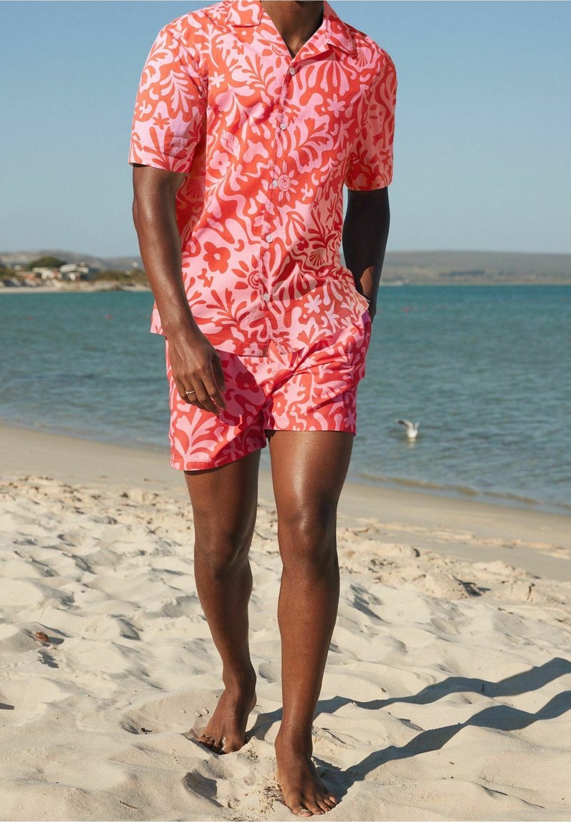 Homme marchant pieds nus sur une plage de sable, portant une chemise à manches courtes florale corail et rose vif et un short assorti.
