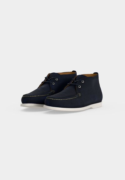 Pair of navy blue leather ankle boots with white soles and black laces on a light gray background.
