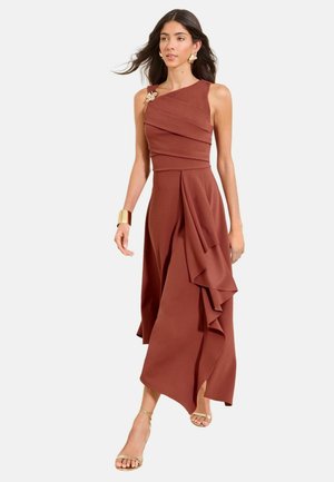 Frau trägt ein ärmelloses, rostfarbenes asymmetrisches Kleid mit Rüschendetails, ein goldenes Manschettenarmband und beigefarbene Riemchenabsätze vor weißem Hintergrund.