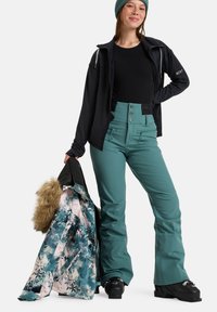 Türkise Skihosen mit hoher Taille, Metallknöpfen und zwei Fronttaschen. Eine schwarze Zip-Jacke und eine farbenfrohe, mit Fellakzenten versehene Skijacke werden gehalten.